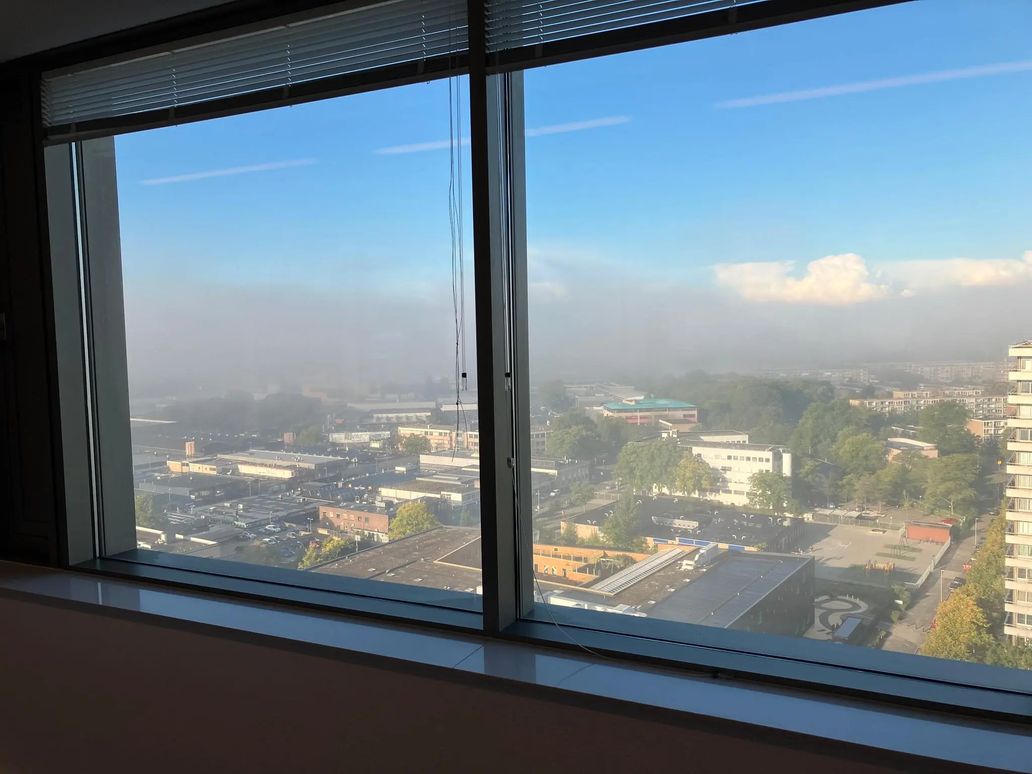 Uitzicht vanuit een hoog kantoorgebouw aan de Vliegend Hertlaan op een mistige ochtend met zicht op een stedelijk gebied en deels heldere lucht.
